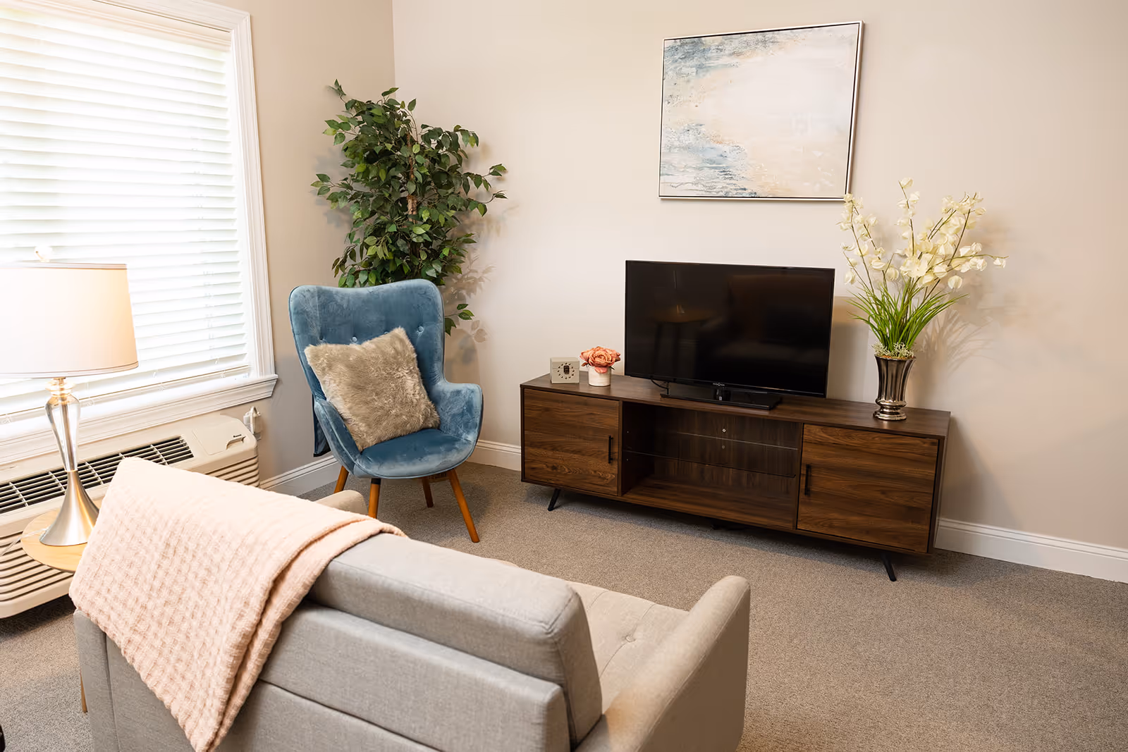 A cozy living room area featuring a light gray sofa with a pink throw blanket draped over the back, a blue upholstered armchair with a furry cushion, a wooden TV stand with a flat-screen television, decorative flowers, and a framed abstract painting on the wall. A large window with white blinds and a table lamp are also visible.