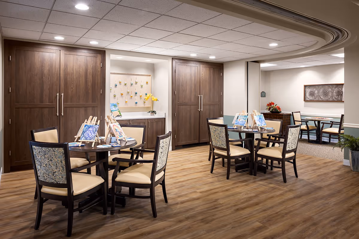 A well-lit interior room with two round tables, each surrounded by four chairs. The tables have small easels displaying paintings and painting supplies. The room features wood flooring, light-colored walls with a green accent, and large wooden cabinets along the back wall. There is a small counter with a flower vase and a bulletin board with hanging items. In the background, another seating area with tables and chairs is visible.