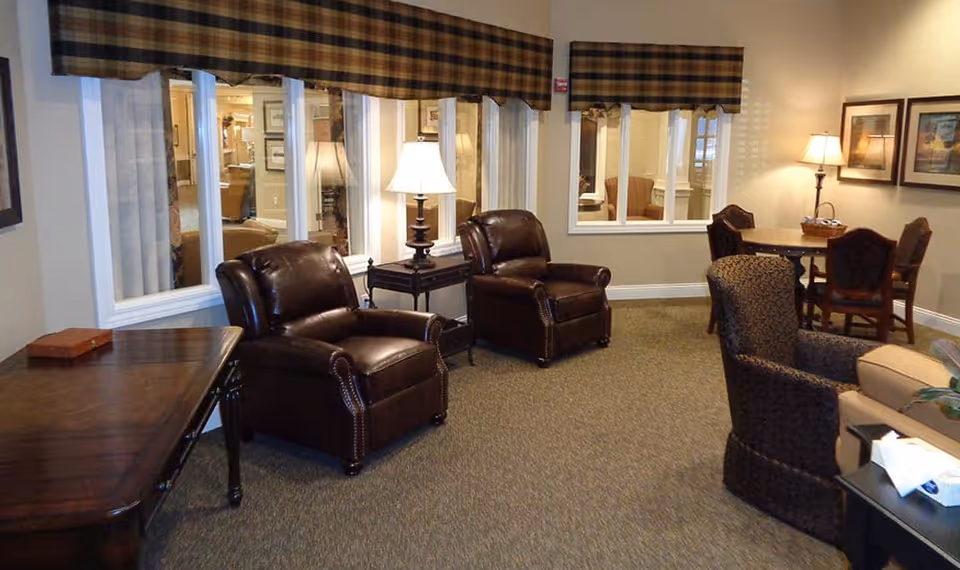 A cozy interior common area with two dark brown leather armchairs, a small table with a lamp between them, a patterned armchair, and a round table with four chairs in the background. The walls are beige with framed artwork and windows with plaid valances.