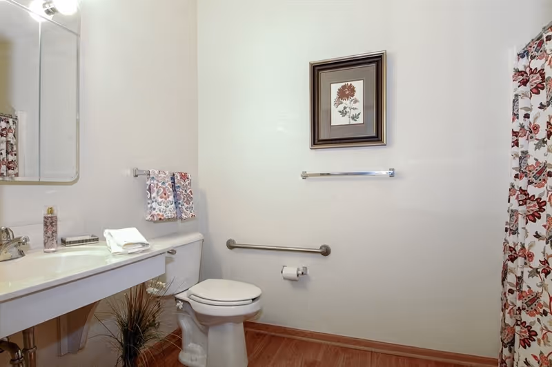 A clean bathroom with a white toilet, a white sink with a silver faucet, a mirror above the sink, a floral hand towel hanging on a rack, a framed flower picture on the wall, a floral shower curtain, and a wooden floor.