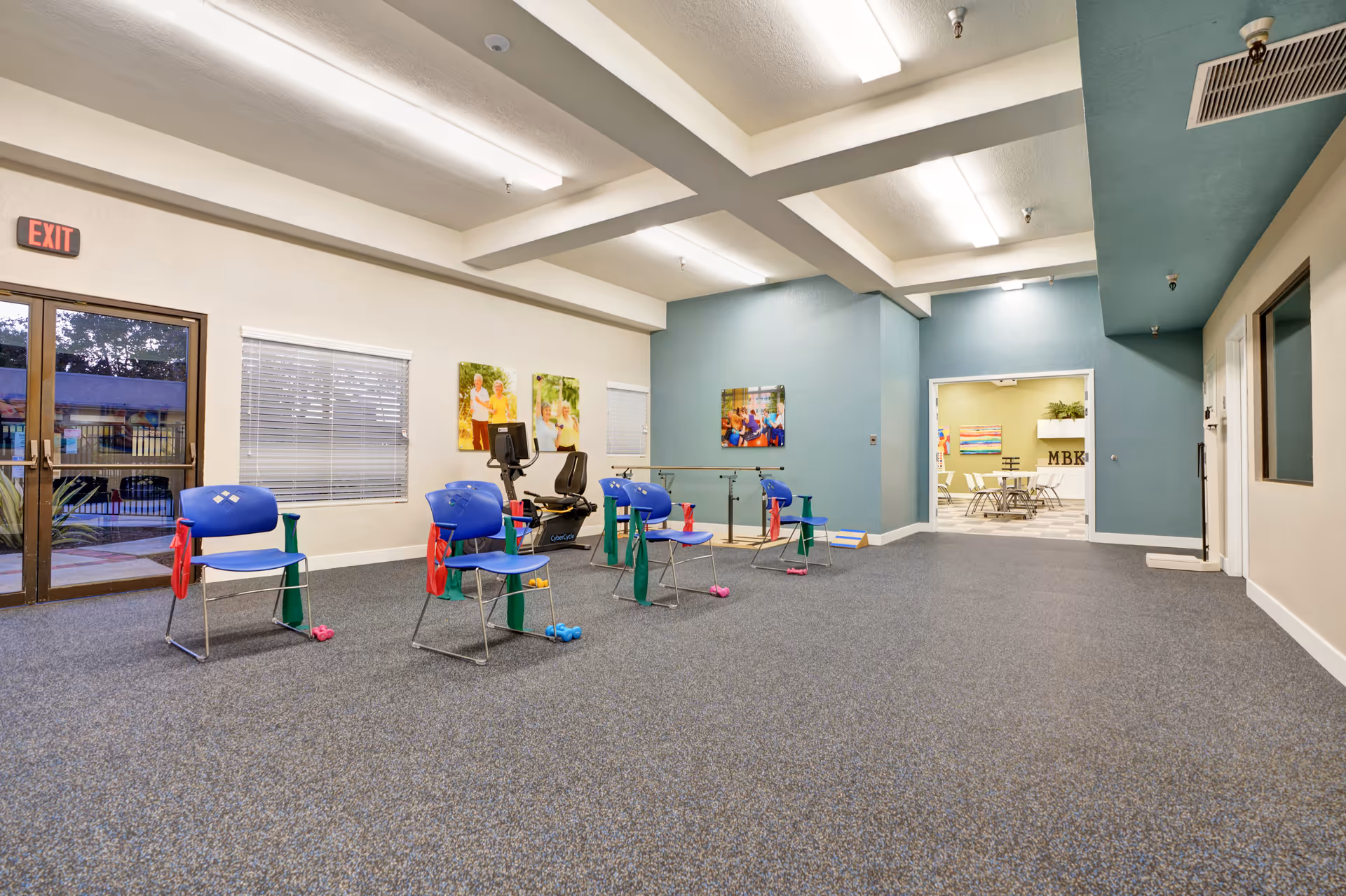 A spacious exercise room in a senior living facility with blue chairs arranged in a circle, each equipped with small hand weights and resistance bands. There is an exercise bike near the window, and colorful pictures of seniors on the walls. The room has a carpeted floor, white ceiling with fluorescent lights, and a door leading to another room with tables and chairs.