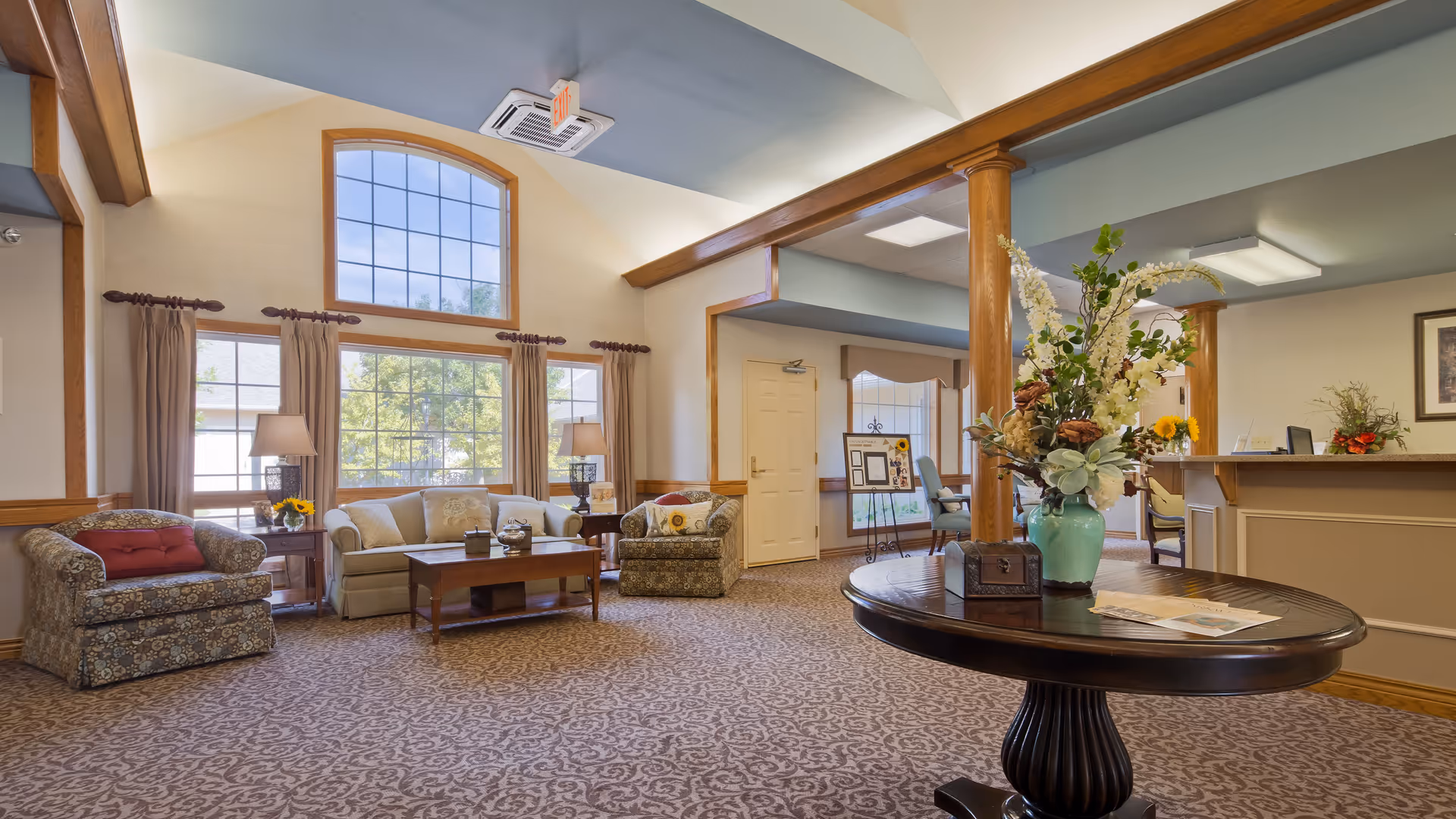 A spacious and well-lit senior living facility common area with large windows, beige walls, and patterned carpet. The room features a seating area with a sofa and two armchairs, a wooden coffee table, and side tables with lamps. In the foreground, there is a round wooden table with a large floral arrangement in a green vase and some papers. The reception desk is visible on the right side with a computer and decorative items. Wooden columns and trim add warmth to the space.