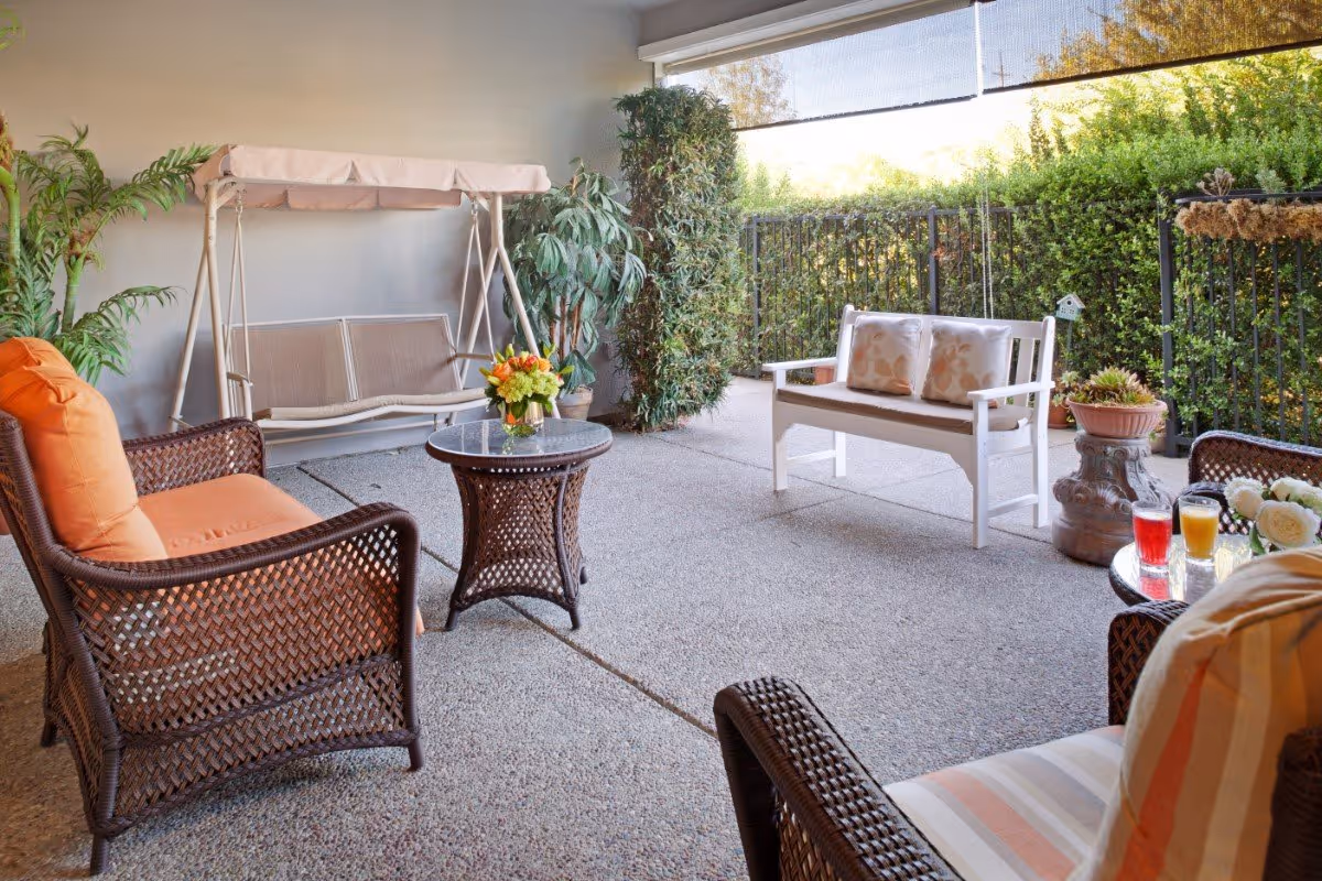A covered outdoor patio area with wicker chairs featuring orange and striped cushions, a white wooden bench with two patterned pillows, a round wicker table with a flower arrangement, and a swing seat with a canopy. The patio is surrounded by greenery and potted plants, with a fence and trees visible in the background.