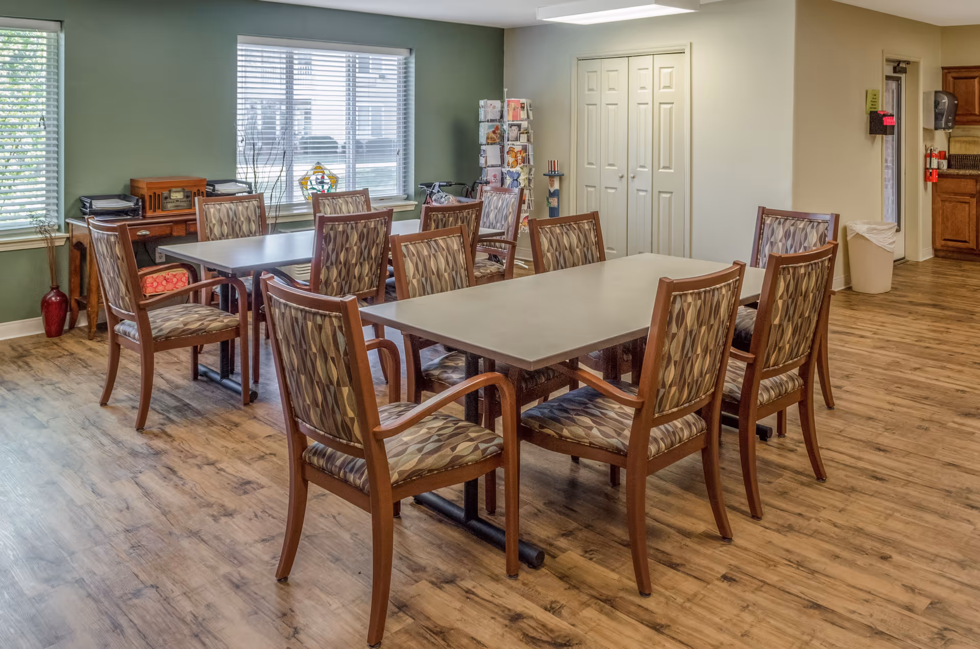 A dining area with two rectangular tables surrounded by wooden chairs with patterned cushions. The room has wood flooring, a green accent wall with two windows, and a small wooden cabinet with decorative items. There is a rack with brochures near a white double-door closet and a kitchen area visible in the background.