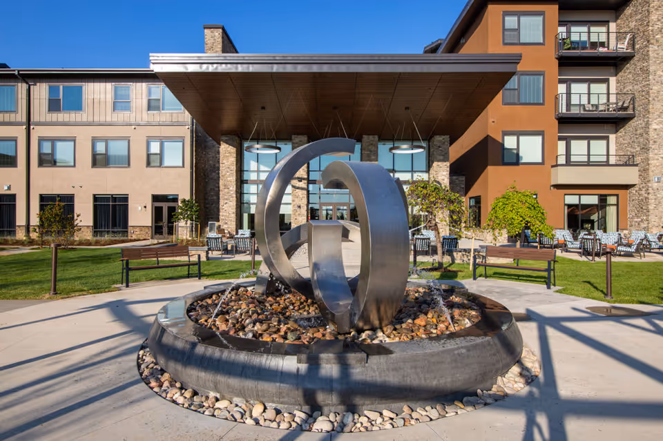 Outdoor view of The Ridge Foothill facility entrance featuring a modern metal sculpture fountain surrounded by rocks and water jets. The building has multiple floors with large windows and balconies, outdoor seating areas with chairs and tables, green lawns, and benches under a clear blue sky.
