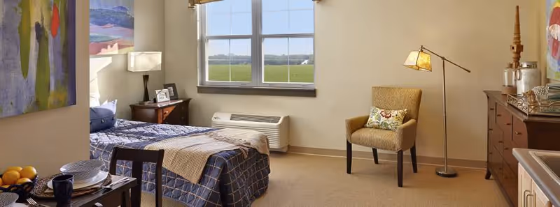 A cozy bedroom in a senior living facility featuring a single bed with blue checkered bedding and a beige throw blanket. There is a wooden nightstand with a lamp and framed photos next to the bed. A window shows a view of green fields outside. The room also includes a cushioned armchair with a decorative pillow, a floor lamp, a wooden dresser with decorative items, and a small dining table set with plates, bowls, and fruit.