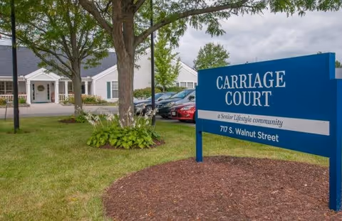 Exterior view of Carriage Court senior living community with a blue sign displaying the name and address, surrounded by green grass, trees, and parked cars near a white building.