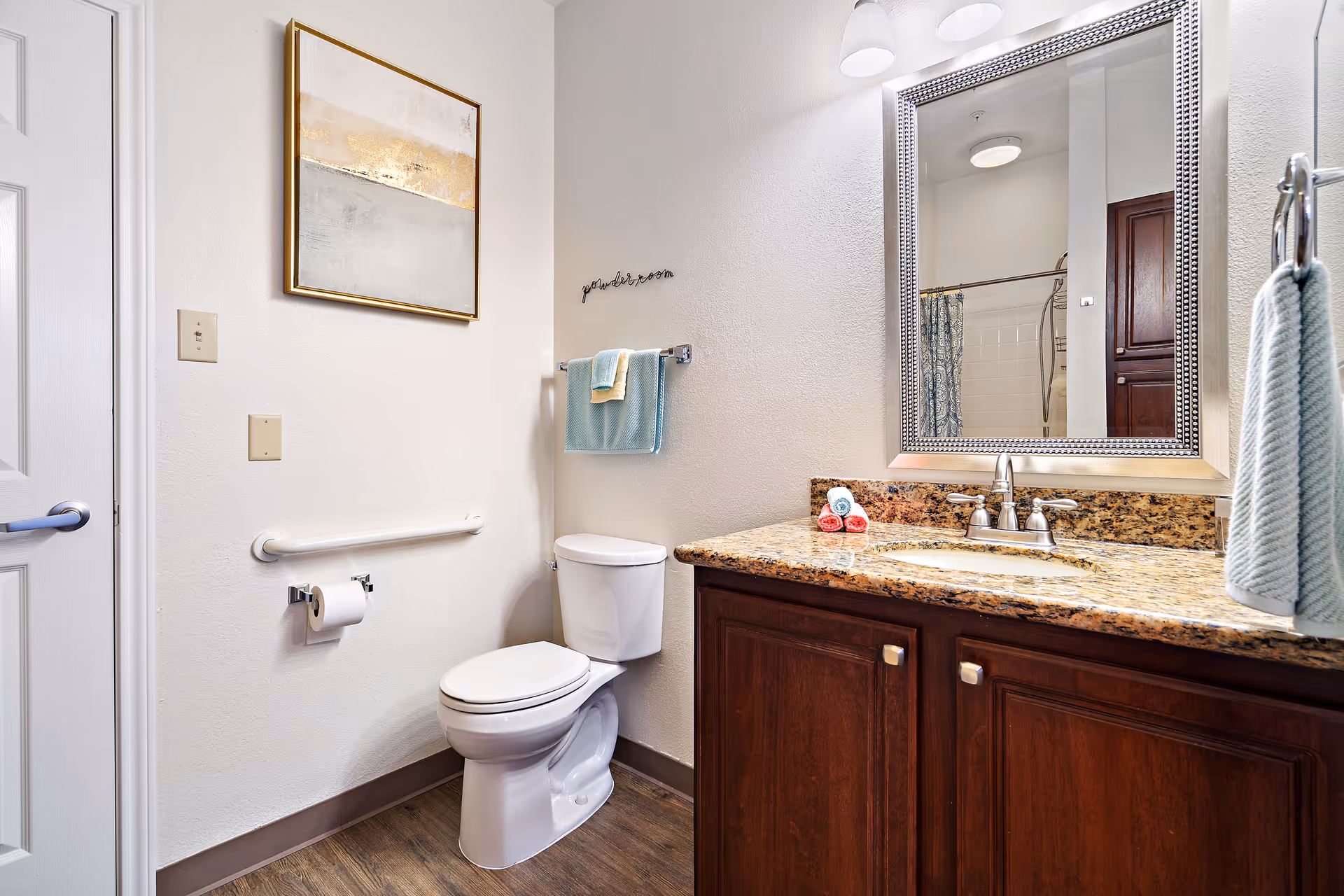 Well-lit bathroom with a toilet, grab bar, granite-topped vanity with mirror, and towels on racks.