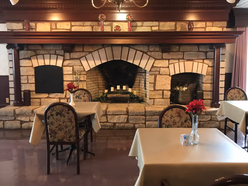 Dining area with tables and floral-upholstered chairs facing a large stone fireplace decorated with candles and flowers.