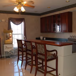 Interior view of a kitchen area with a wooden countertop bar and three high chairs with patterned cushions. There is a ceiling fan with lights above, a window with a curtain, a popcorn machine on the left side, and wooden cabinets mounted on the wall.