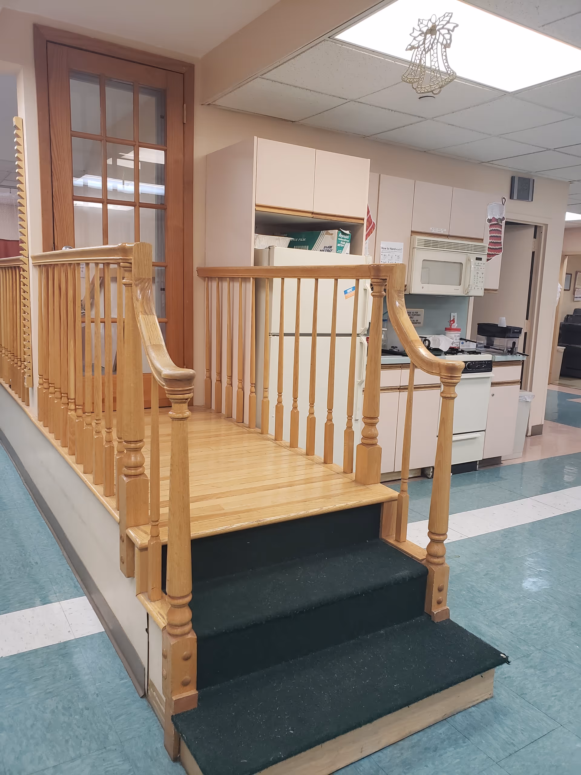 Small raised wooden platform with spindle railings and carpeted steps leading up to a kitchenette area with a refrigerator and microwave.