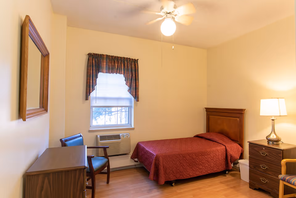 A small, simple bedroom with a single bed covered in a red quilt, a wooden headboard, a wooden dresser with a lamp on it, a chair with a blue cushion, a window with blinds and a plaid valance, a wall-mounted mirror, and a ceiling fan with a light.