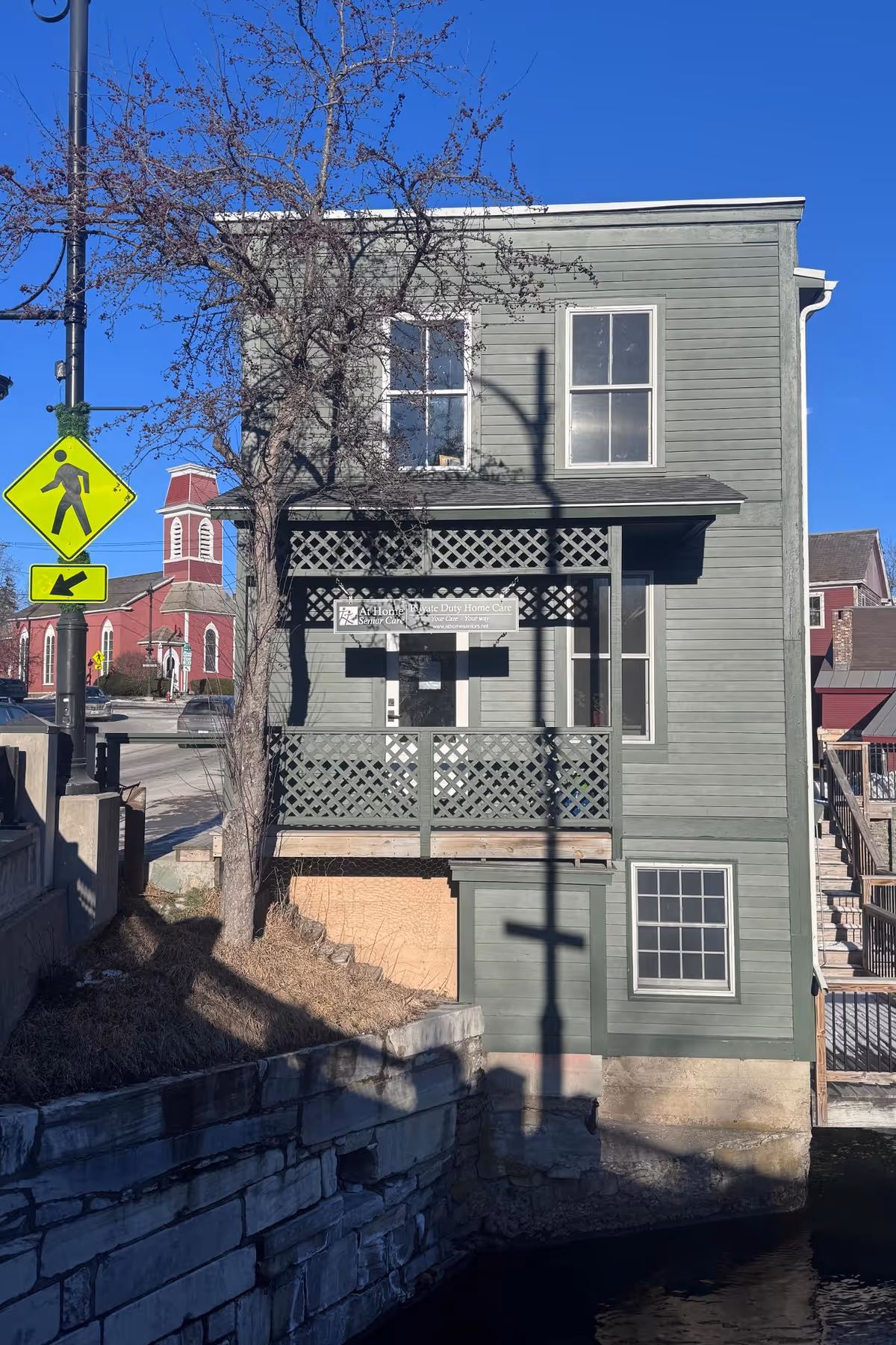 Two-story green wooden building with a lattice porch displaying an 'At Home Senior Care' sign, next to a pedestrian crossing sign.