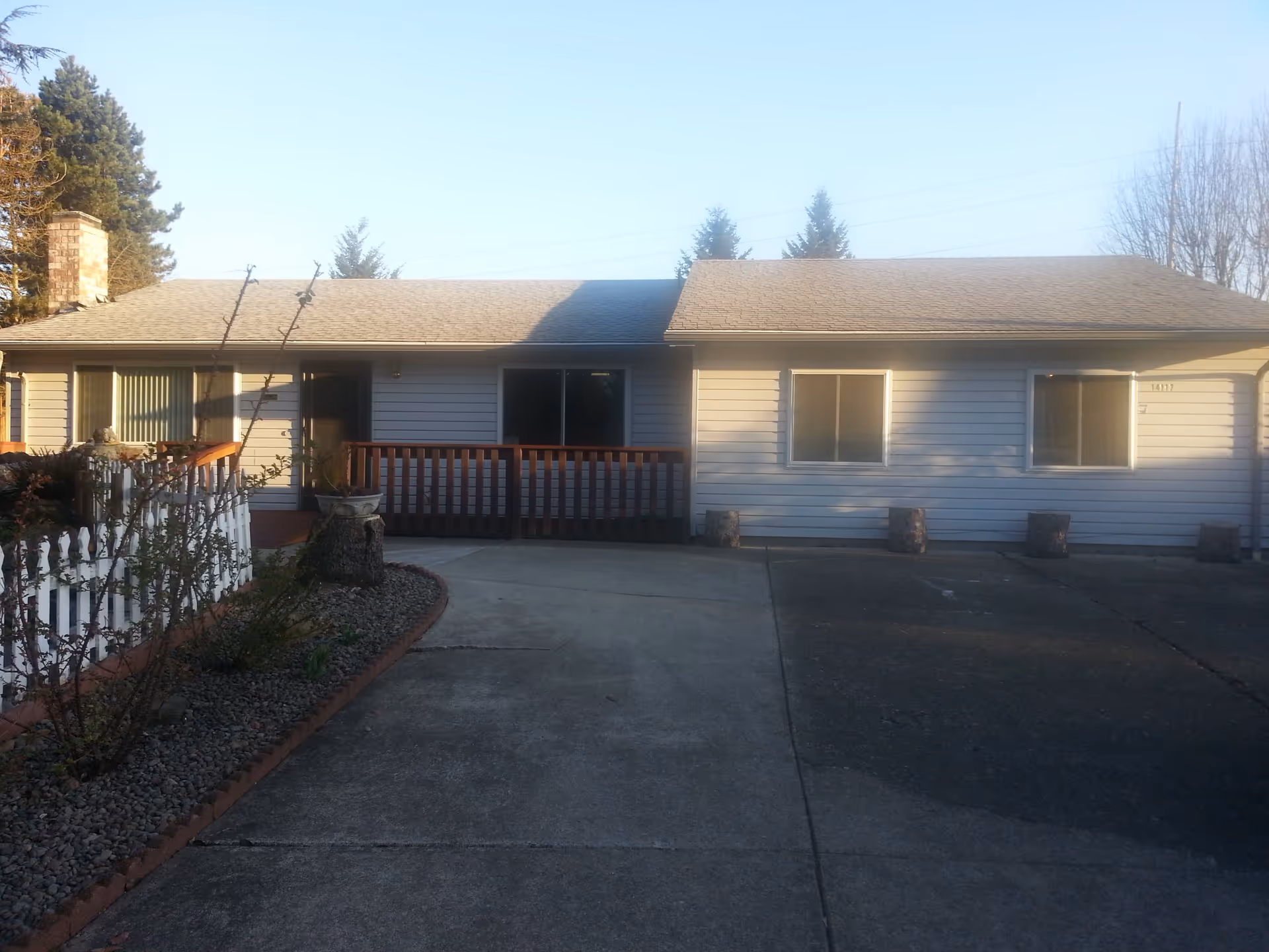 Front exterior view of a single-story residential building with light-colored siding, a shingled roof, and several windows. There is a concrete driveway leading up to the house, a small garden area with plants and a white picket fence on the left side, and a wooden railing near the entrance. Trees are visible in the background under a clear sky.