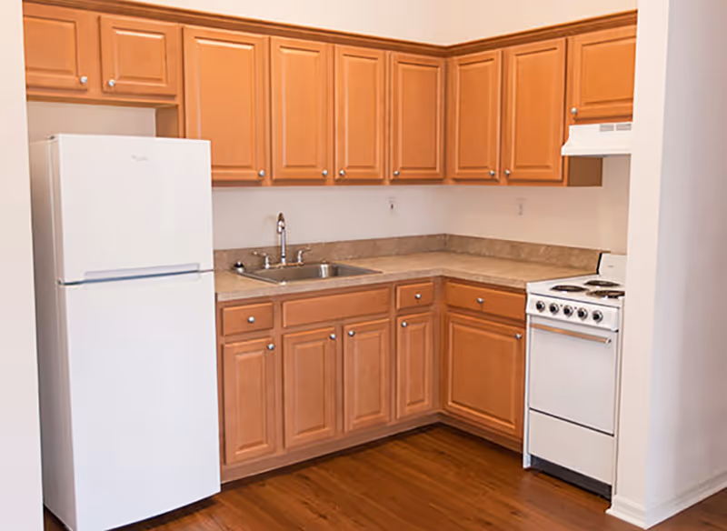 A small kitchen with wooden cabinets, a white refrigerator, a stainless steel sink, and a white electric stove with an oven. The floor is wooden and the walls are painted white.