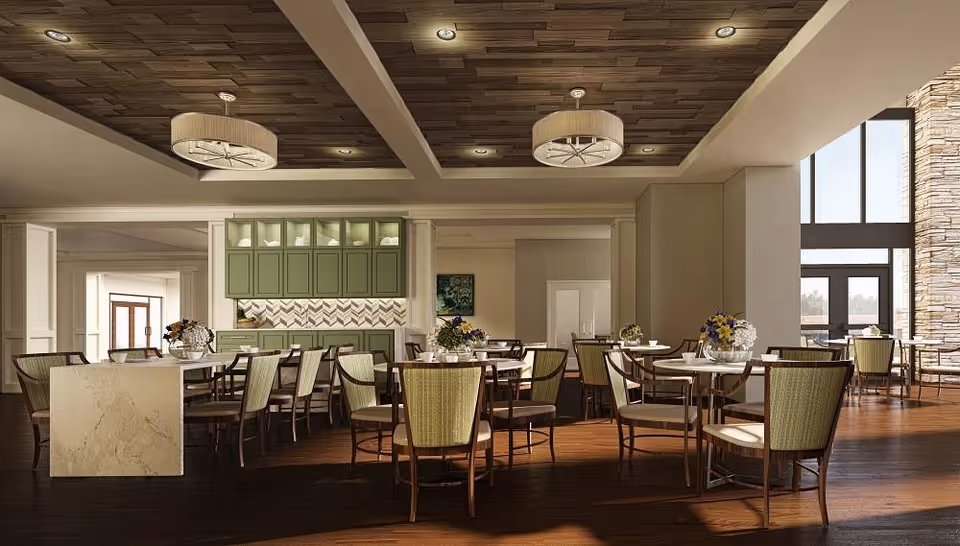 A spacious and well-lit dining room with multiple round tables and chairs arranged neatly. The room features a wooden ceiling with recessed lighting and two large decorative ceiling lights. There are green cabinets and a countertop area along one wall, with floral centerpieces on the tables. Large windows on the right side allow natural light to enter, highlighting the warm wooden flooring and stone accent wall near the entrance.
