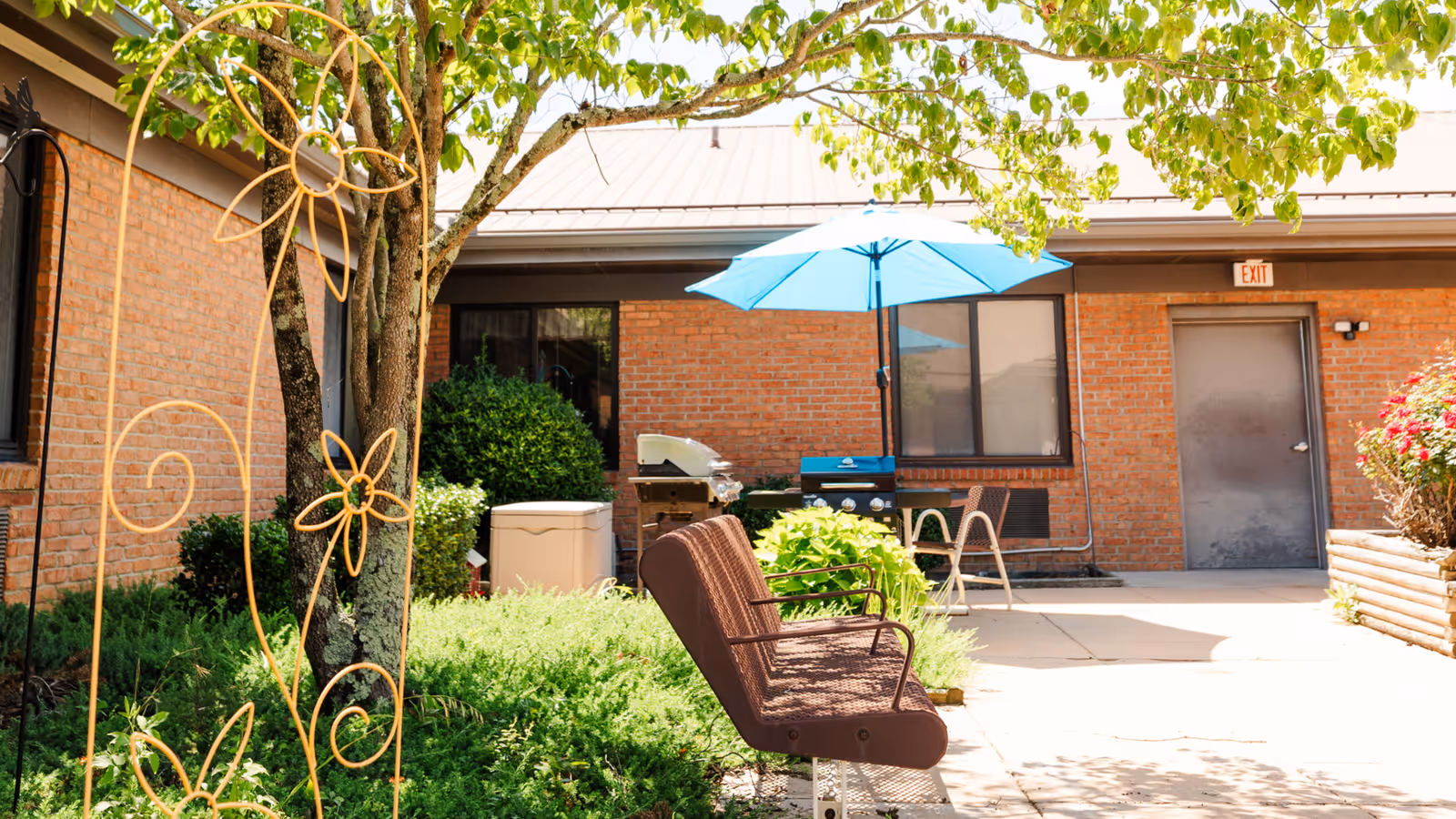 Outdoor courtyard area with a brown bench, a blue patio umbrella, a tree with green leaves, decorative metal flowers, bushes, and a brick building with windows and a door in the background.