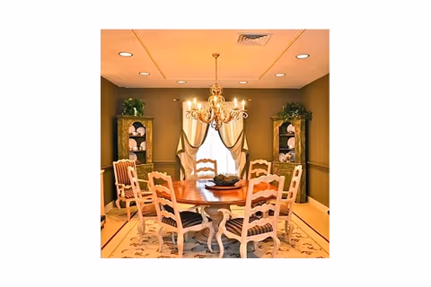 A formal dining room with a round wooden table surrounded by eight white chairs with striped cushions. The room features a chandelier hanging from the ceiling, two tall cabinets with decorative plates, and a window with beige curtains. The walls are painted in a warm brown color and the floor has a patterned rug.