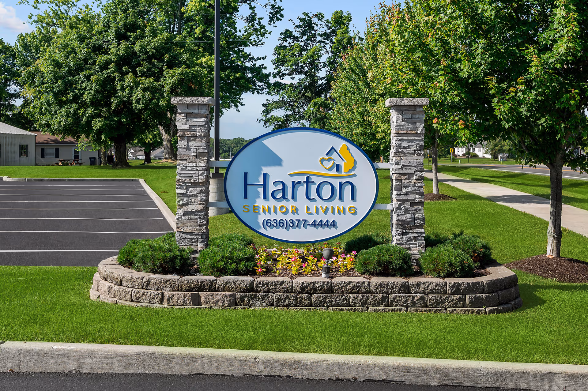 A landscaped entrance sign reading "Harton Senior Living" between stone pillars with a parking lot, sidewalk and trees behind it.