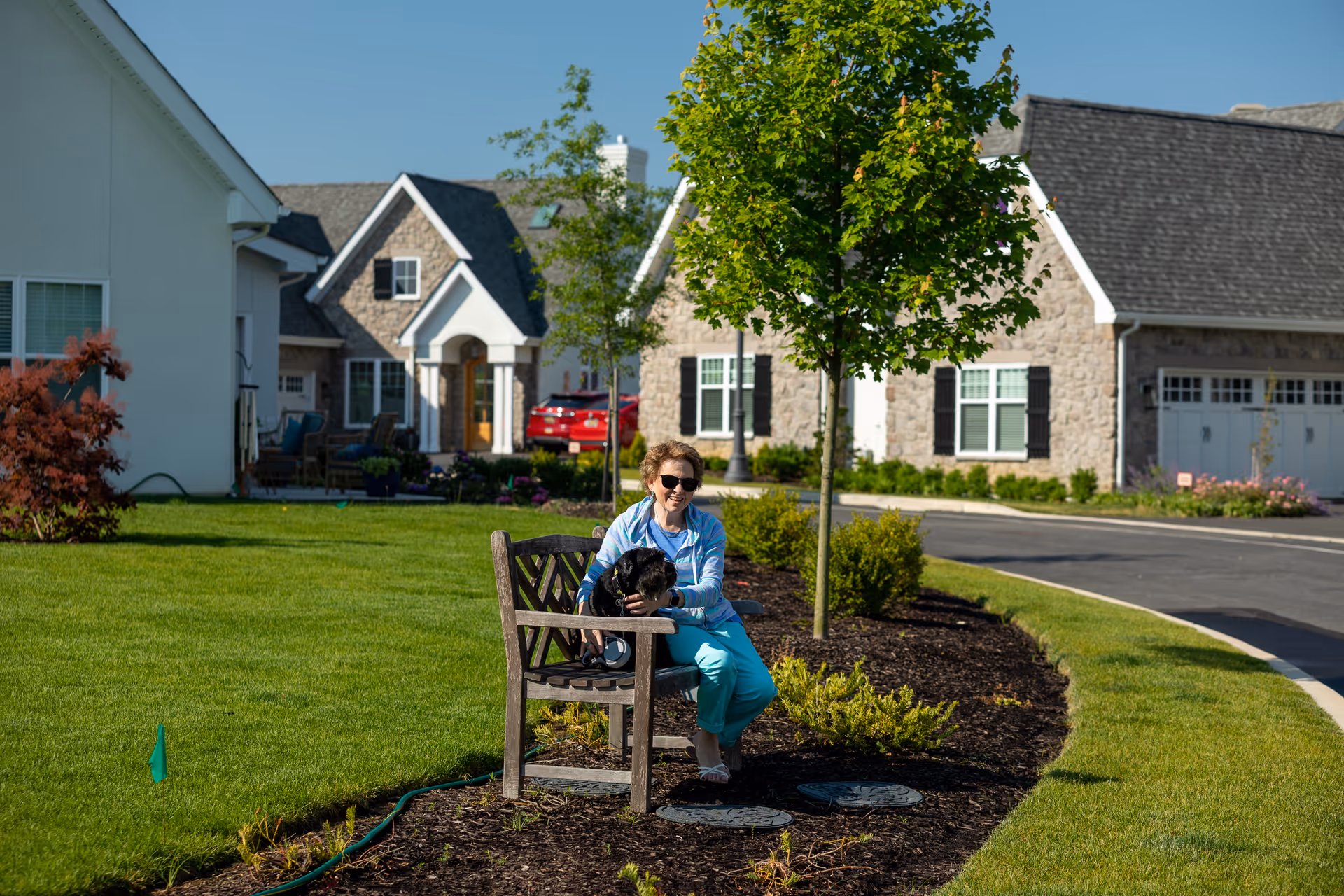 A woman wearing sunglasses and blue clothing sits on a wooden bench in a landscaped garden area with a small black dog on her lap. Behind her are residential-style buildings with stone and white siding, a driveway, and a red car parked near the entrance. The sky is clear and blue.