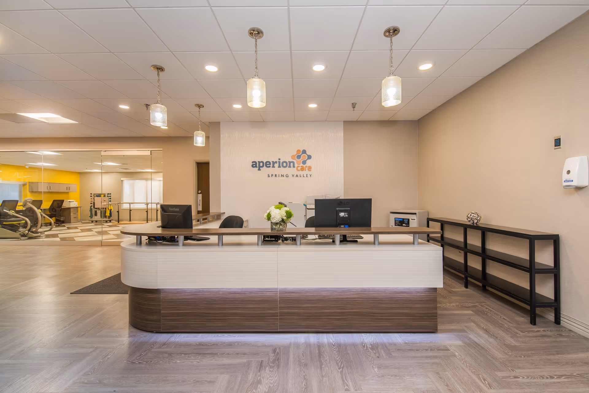Reception area of a senior living facility with a modern curved desk, two computer monitors, a flower arrangement, and pendant lights hanging from the ceiling. Behind the desk is a wall with the logo 'aperion care SPRING VALLEY'. To the left, there is a glass wall revealing a room with exercise equipment and a bright yellow accent wall.