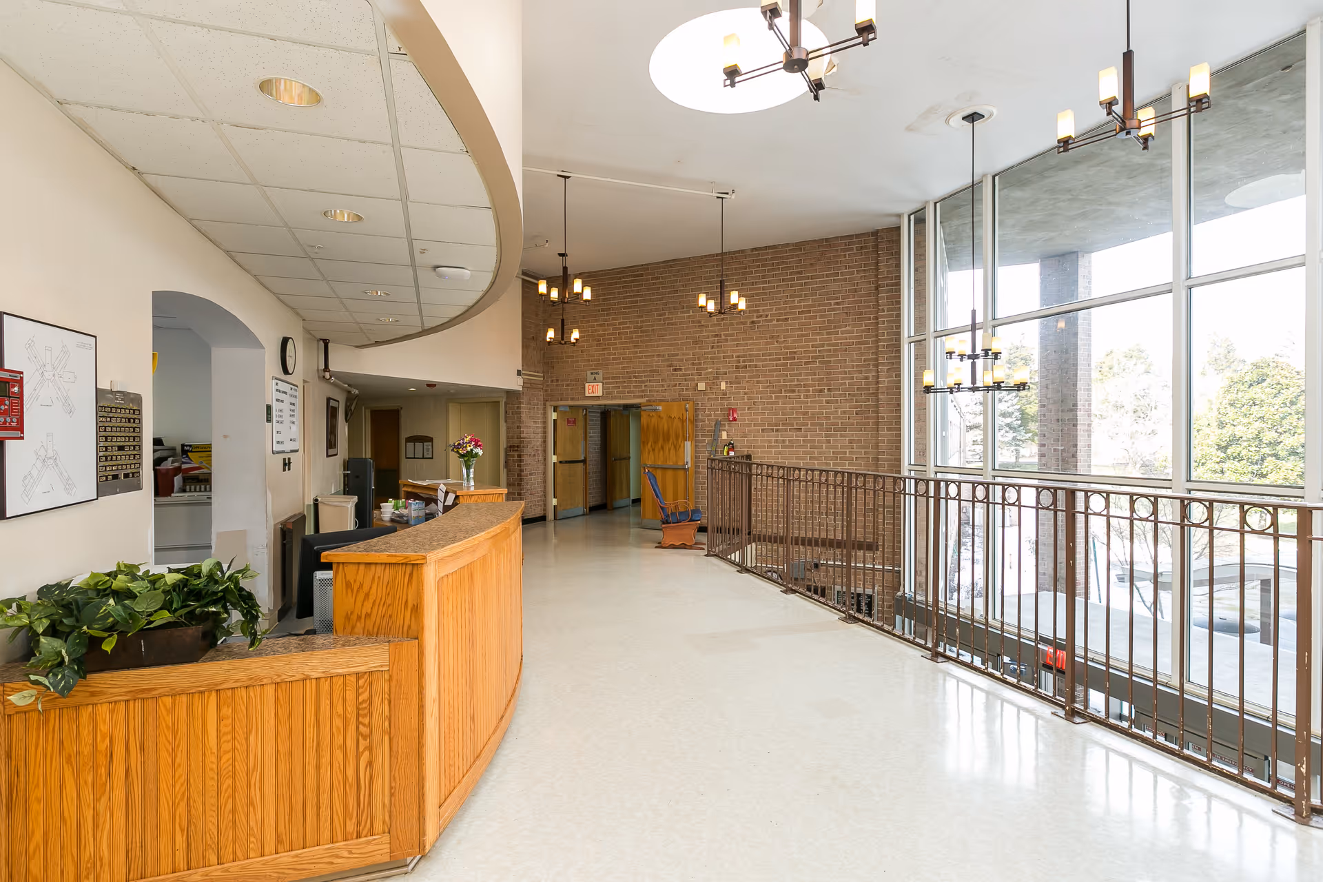 Interior view of a nursing and rehabilitation facility reception area with a wooden front desk, potted plants, a wall-mounted map, and large windows letting in natural light. The space has a high ceiling with hanging light fixtures and a railing overlooking a lower level.