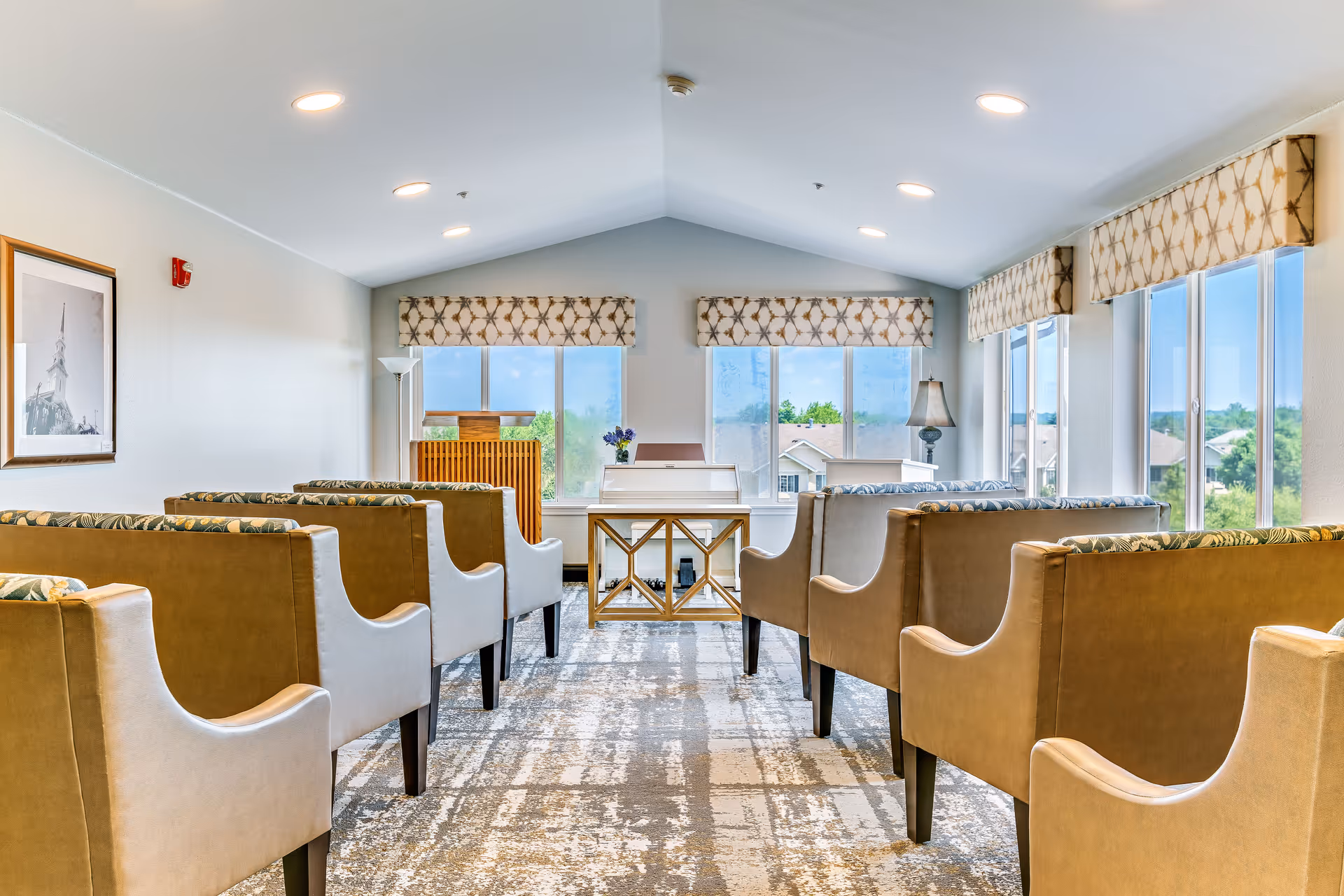 A bright and airy room with rows of beige upholstered chairs facing a white piano and a wooden podium. Large windows with patterned valances let in natural light and offer views of rooftops and greenery outside. The room has a high ceiling with recessed lighting and a patterned carpet.
