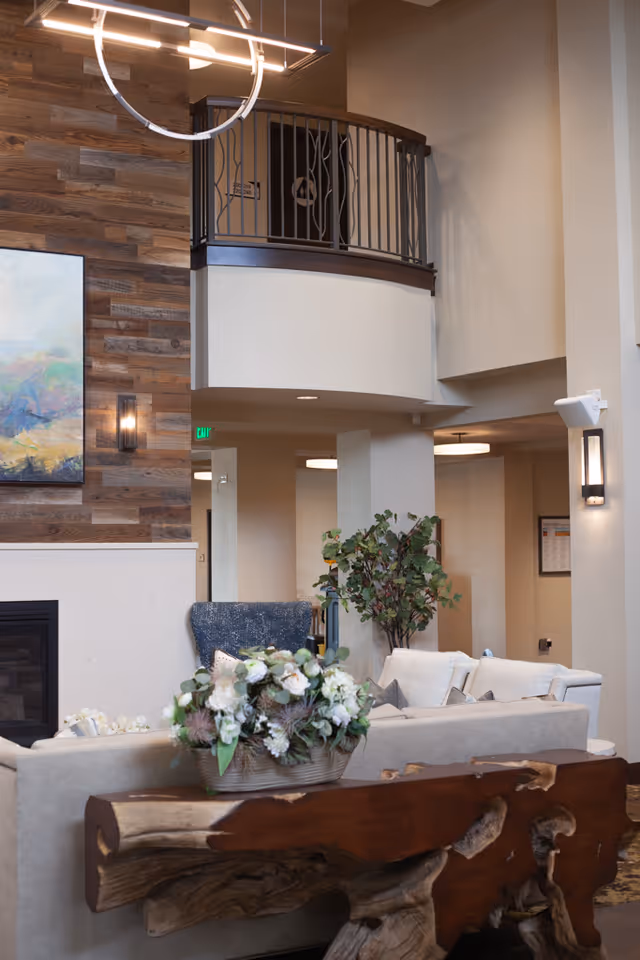 Spacious senior living lounge with sofas, a floral centerpiece on a rustic wooden table, balcony railing and modern lighting.