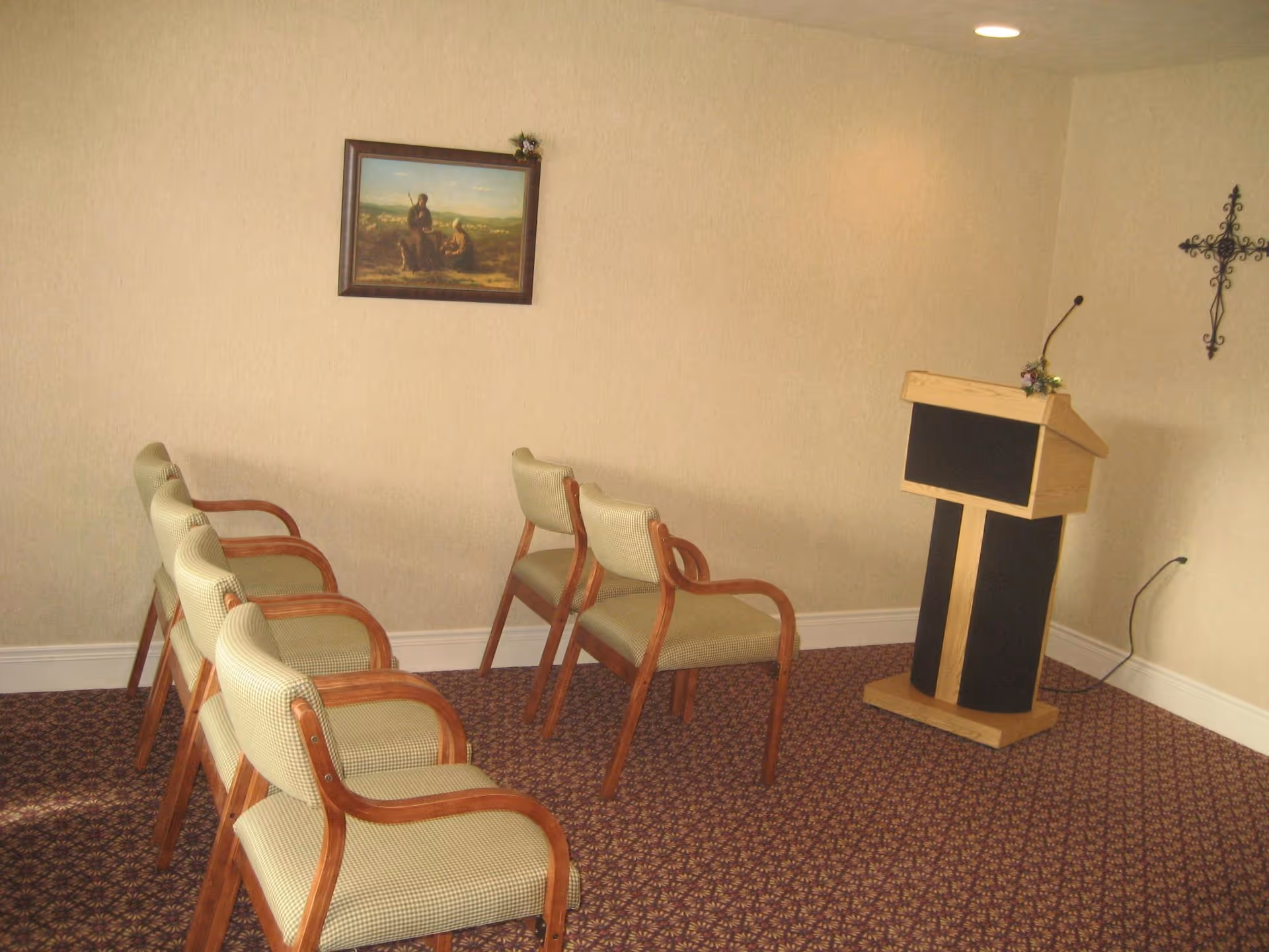 Small meeting or presentation room with six wooden chairs with light green upholstery arranged in two rows facing a wooden podium with a microphone. The room has beige walls, a patterned carpet, a framed painting on the wall, and a decorative metal cross.