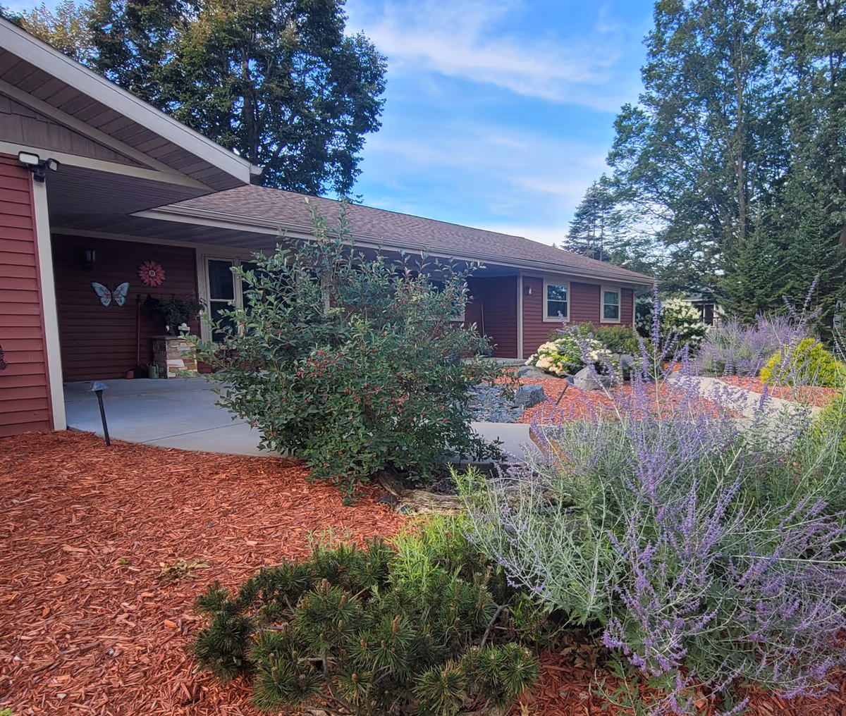 Front exterior of a single-story red assisted-living building with a covered entrance, concrete walkway, and landscaped beds of shrubs, purple flowers, and red mulch.