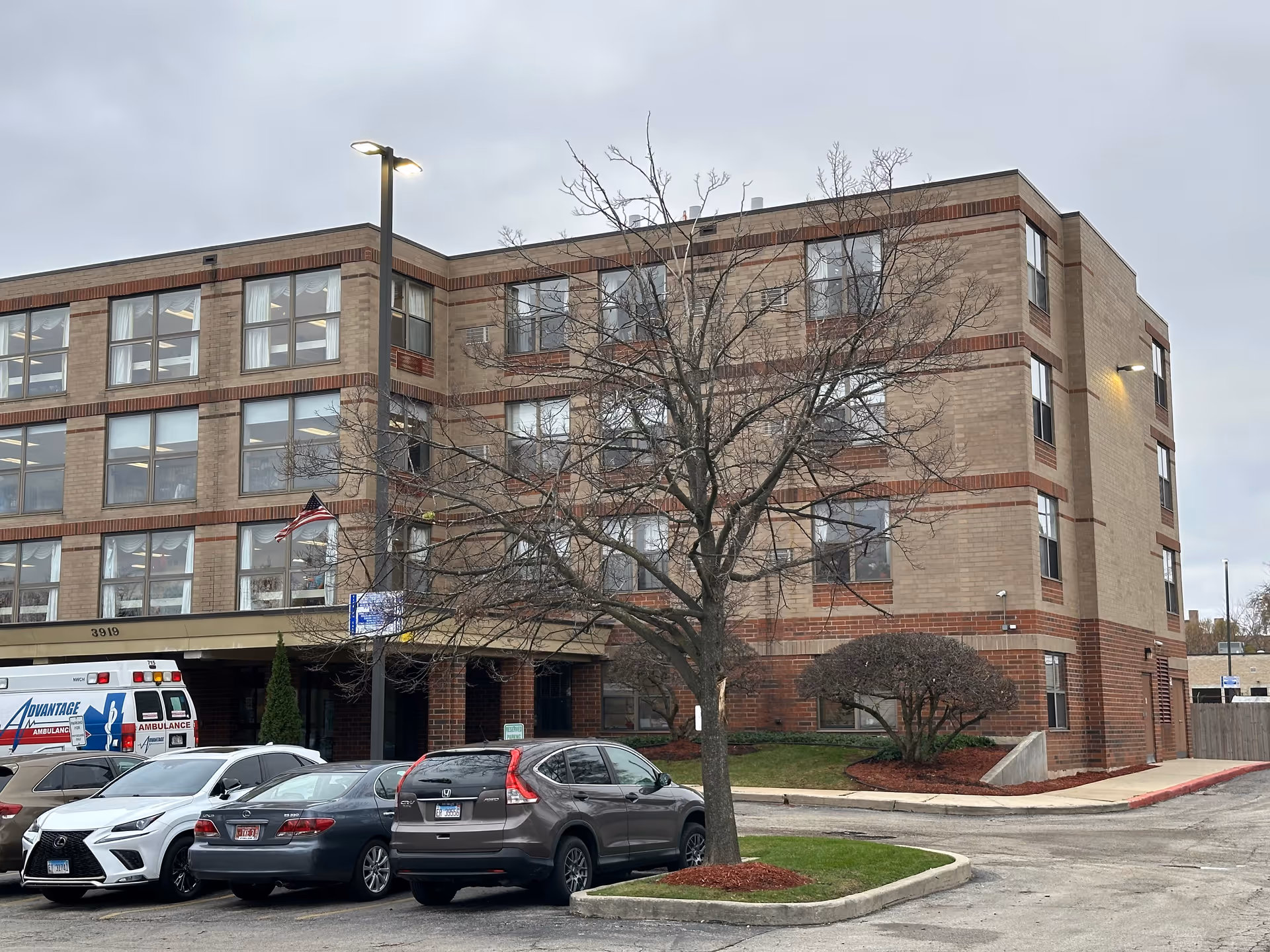 Exterior view of a multi-story brick building with several windows, a leafless tree in front, parked cars, and an ambulance. The sky is overcast.
