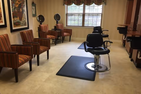 Interior of a salon room with two black salon chairs on mats in front of sinks and mirrors, four striped armchairs along the left wall, framed artwork on the walls, and a window with blinds and a valance letting in natural light.