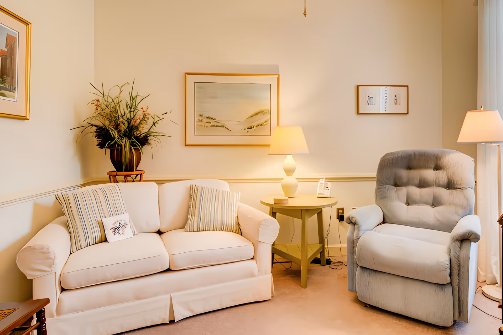 A cozy living room with a white sofa adorned with striped pillows, a light gray cushioned recliner chair, a round wooden side table with a white lamp, and framed artwork on the walls. A potted plant is placed on a stand in the corner.