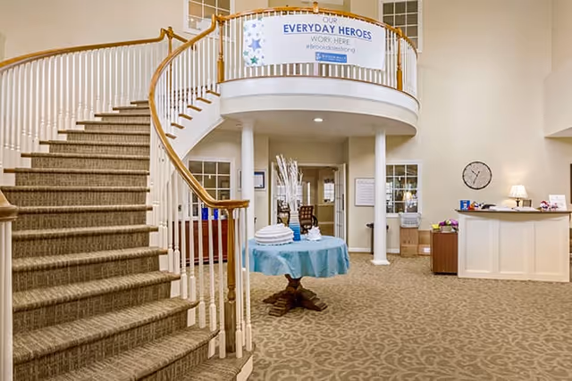 Interior view of a senior living facility lobby with a curved staircase on the left, a round table with a blue tablecloth and decorative items in the center, and a reception desk on the right. A banner above the entrance reads 'Our Everyday Heroes Work Here #BrookdaleStrong'.