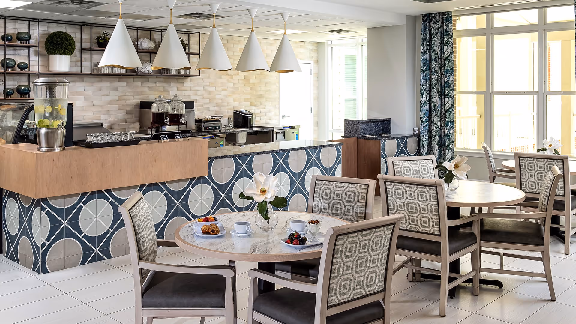 A bright and modern dining area with round tables and cushioned chairs featuring patterned upholstery. Each table has a small vase with a white flower. The background shows a counter with a geometric tile design, pendant lights hanging above, and beverage dispensers. Large windows with floral curtains allow natural light to fill the space.