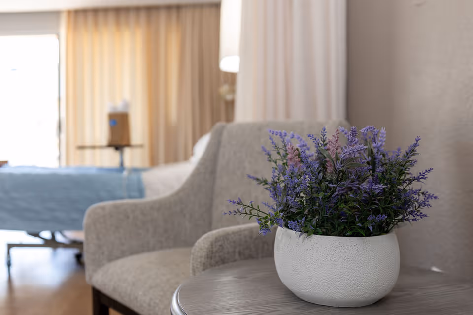 A close-up of a white textured pot with purple and green artificial flowers placed on a round wooden table. In the background, there is a beige upholstered armchair and a hospital-style bed with a blue blanket, partially visible in a softly lit room with beige curtains.