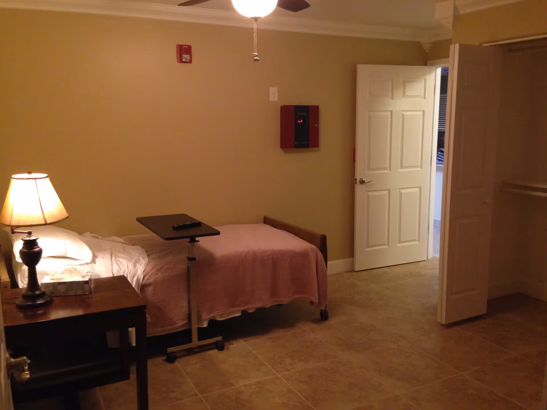 Furnished bedroom with a single bed covered by a pink blanket, bedside table and lamp, an overbed table, and an open door to a hallway.