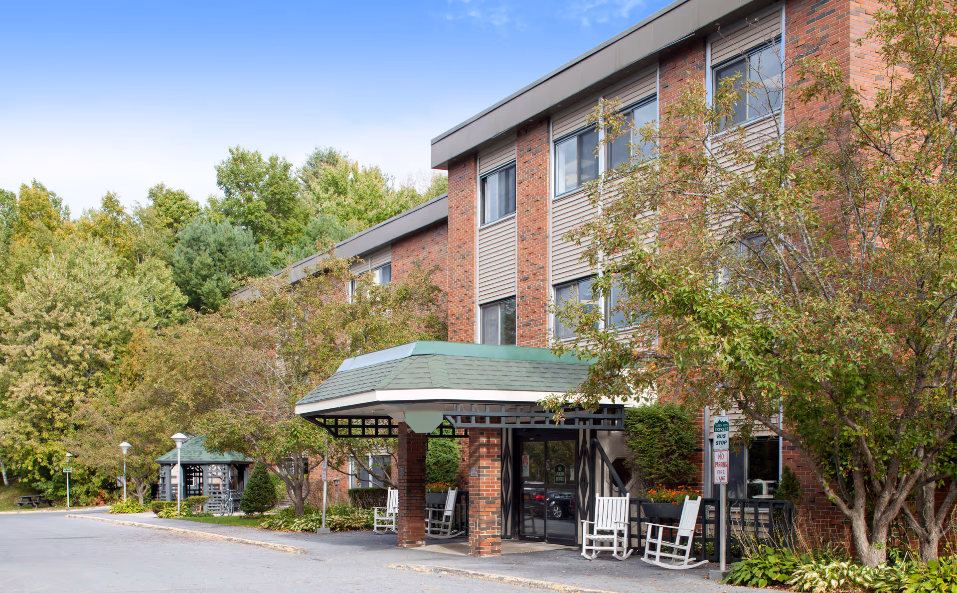 Front entrance of a brick senior living building with a covered porte-cochère, rocking chairs, and surrounding trees.