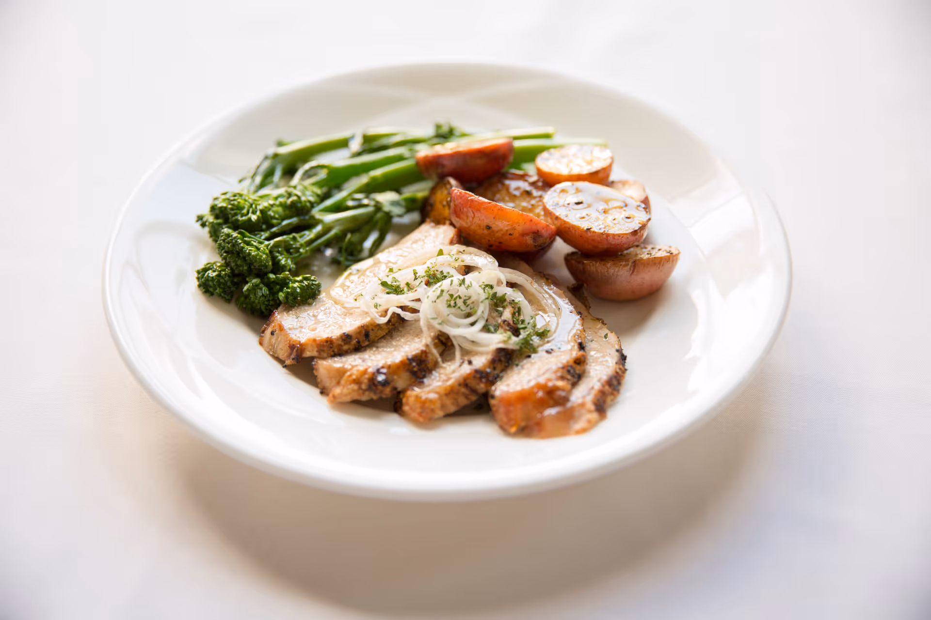 A white plate with a meal consisting of sliced roasted meat topped with onions and herbs, roasted baby potatoes, and steamed broccolini, placed on a white tablecloth.