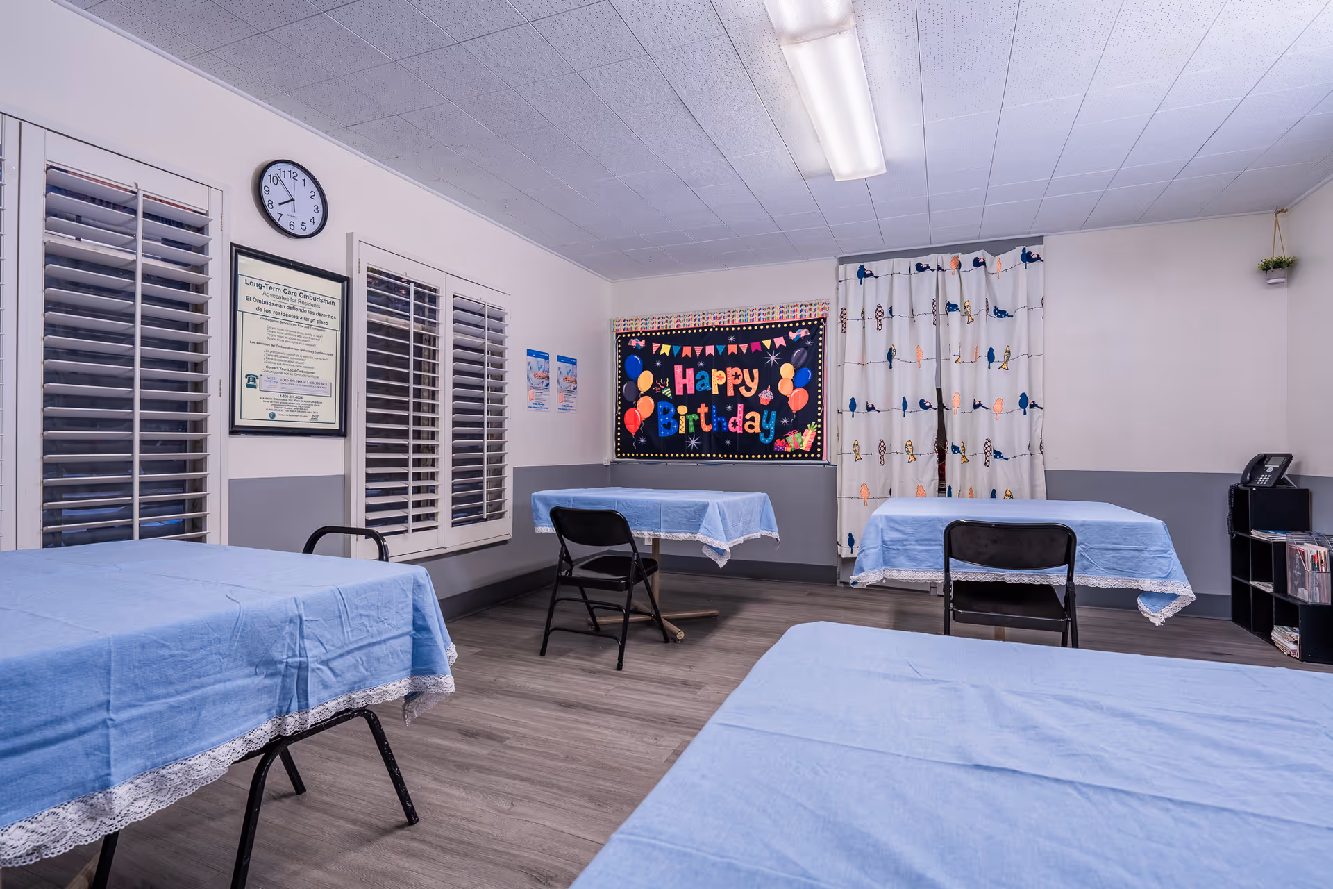 Communal room with several tables covered in blue tablecloths, folding chairs, and a colorful 'Happy Birthday' bulletin board on the wall.