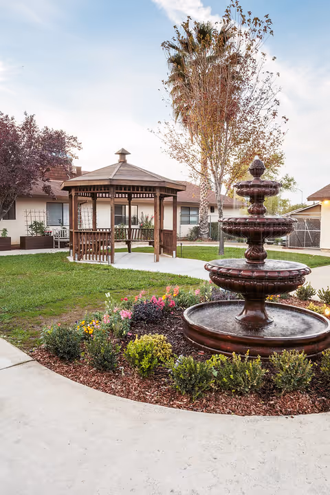 Outdoor garden area at Lincoln Meadows Care Center featuring a three-tiered water fountain surrounded by flowers and shrubs, a wooden gazebo with benches, and trees with autumn foliage under a partly cloudy sky.