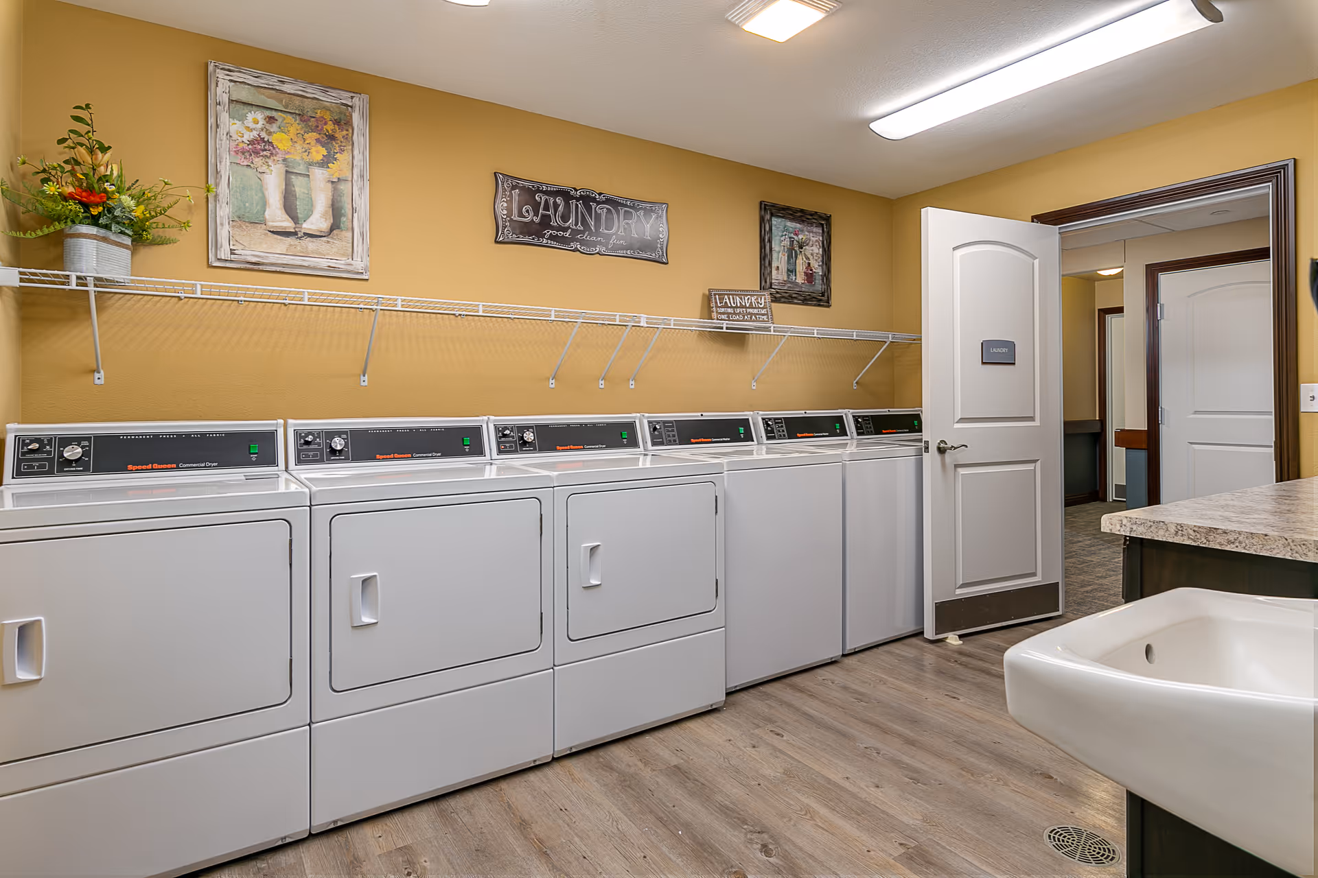 Laundry room with multiple white commercial washers and dryers lined up against a mustard yellow wall. Above the machines is a white wire shelf with decorative items including a flower arrangement and framed pictures. A door labeled 'Laundry' is open, showing a hallway beyond. The floor is wood-style laminate and there is a white utility sink in the foreground.