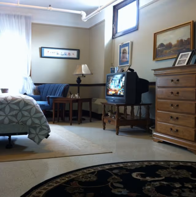 A cozy room with a bed partially visible on the left, a blue upholstered armchair, a wooden side table with a lamp, an old-style CRT television on a wooden stand, a wooden dresser, framed pictures on the walls, and a patterned rug on the floor.