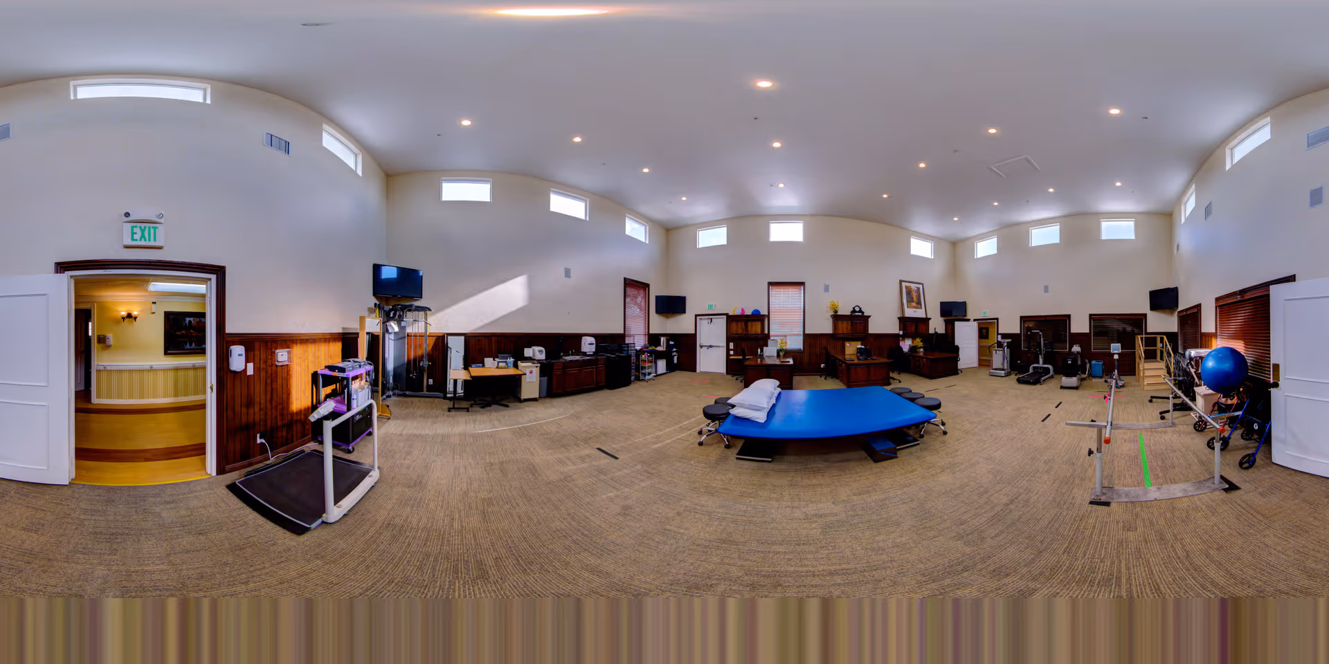 Wide, well-lit interior therapy room with a central treatment table, treadmills, parallel bars, exercise equipment and cabinetry under high clerestory windows.