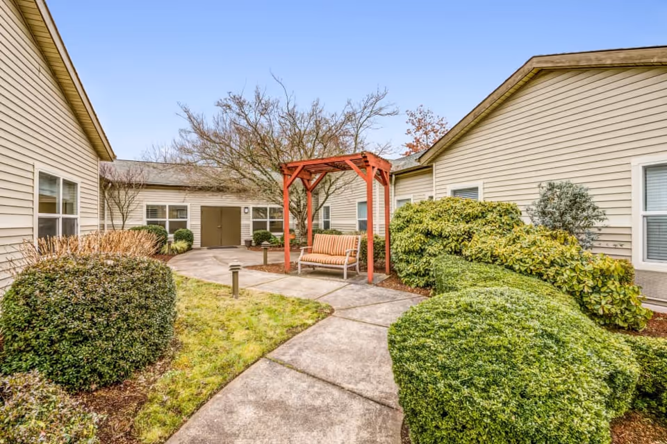 Courtyard between beige single-story buildings with a bench under a red wooden pergola and landscaped shrubs and a paved walkway.