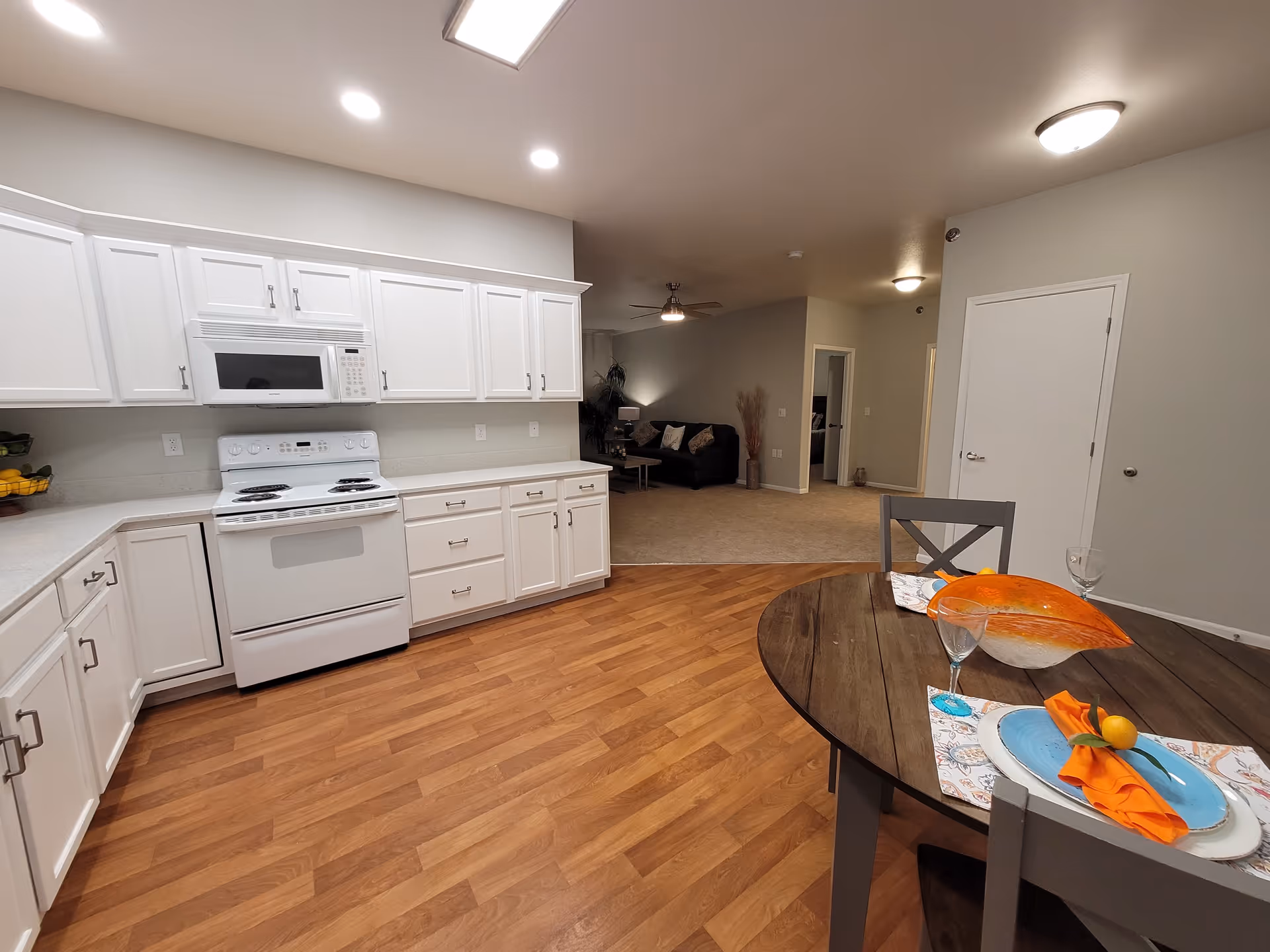 Open-plan kitchen with white cabinets and stove, a small round dining table set in the foreground, and a living area visible beyond.