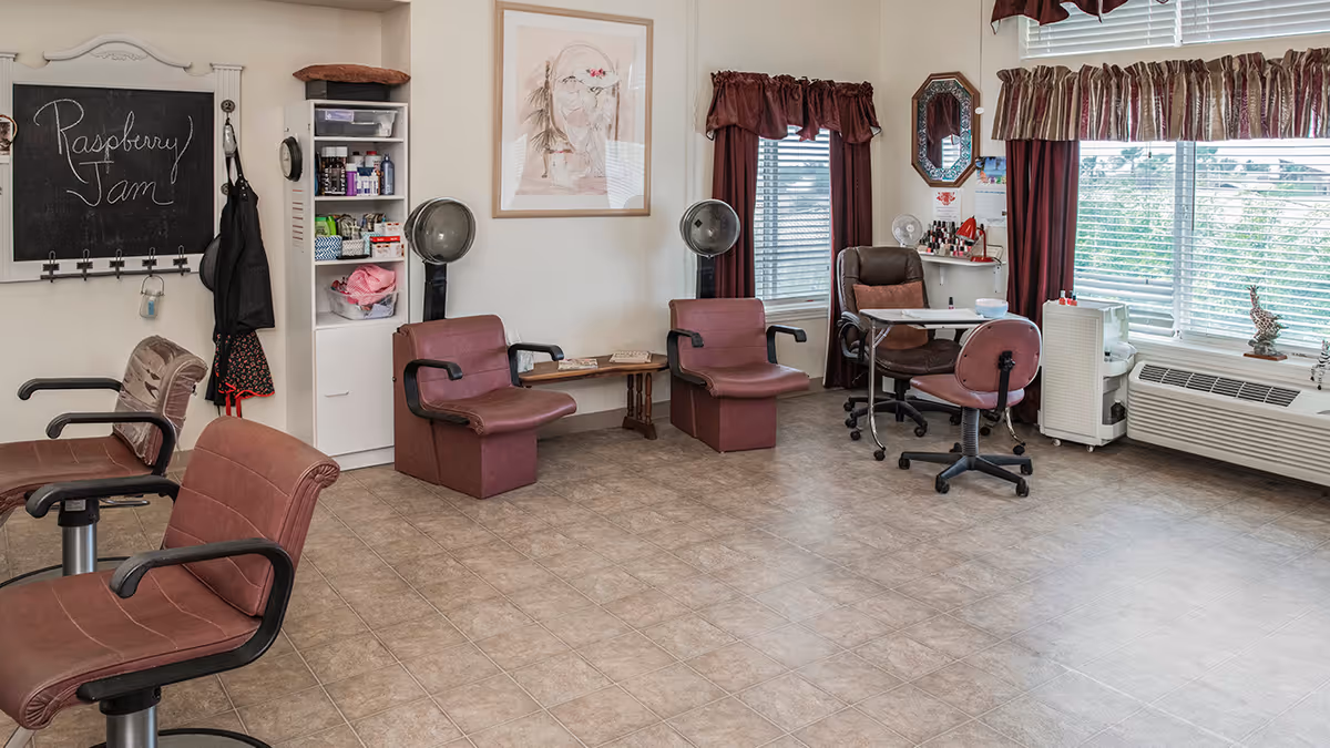 Interior of a salon area in a senior living facility with several brown salon chairs, two hair dryer chairs, a small table with nail polish and manicure tools, windows with maroon curtains, and a chalkboard on the wall with the words 'Raspberry Jam' written on it.