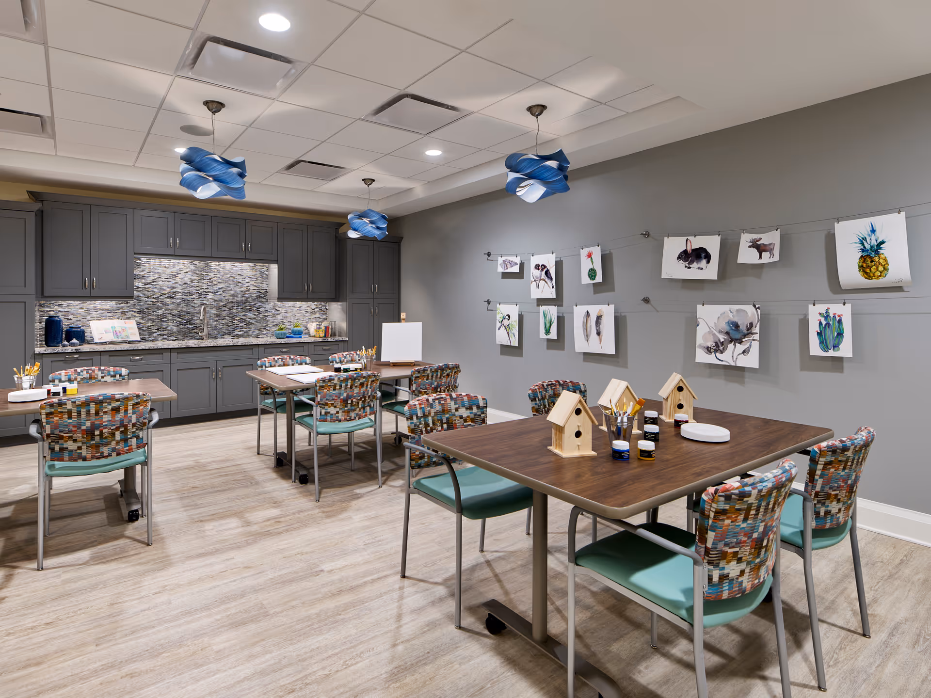 Activity/craft room with tables and colorful chairs, a kitchenette along the back, and artwork displayed on the wall.