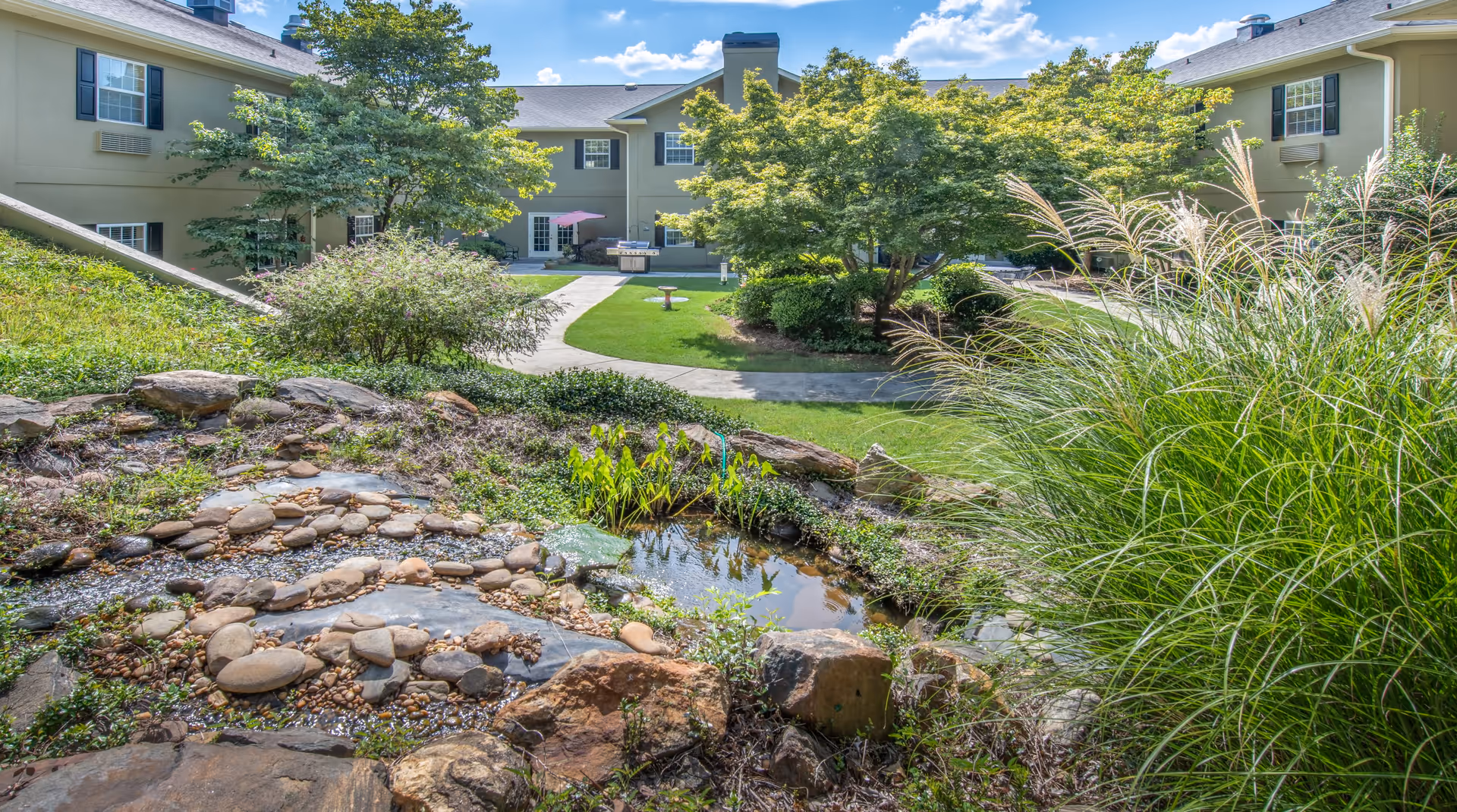 Landscaped courtyard with a small rock-lined pond, walking paths, trees and grassy areas surrounded by a two-story senior living building.