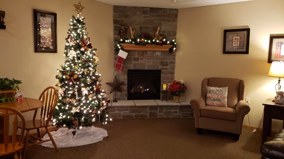 Cozy living room decorated for Christmas with a lit Christmas tree, a stone fireplace adorned with garland and a stocking, a comfortable armchair with a festive pillow, a wooden dining table with chairs, and warm lighting from a table lamp.